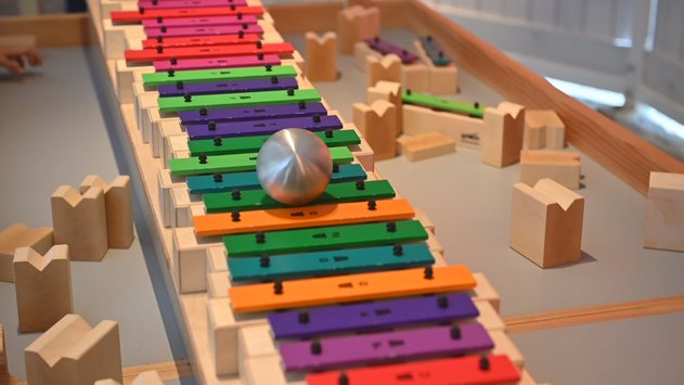 Ein Klangxylophon, das aus bunten, aneinandergereihten Metallplättchen besteht.  Auf diesen Klangplatten rollte gerade eine Metallkugel entlang.